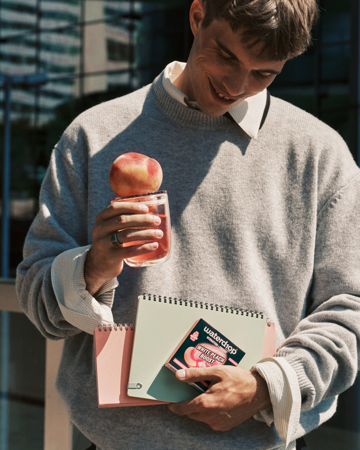 Man in grijs pullover staat buiten en houdt een roze drankje met een appel aan de rand vast, terwijl hij een notitieboek vasthoudt.