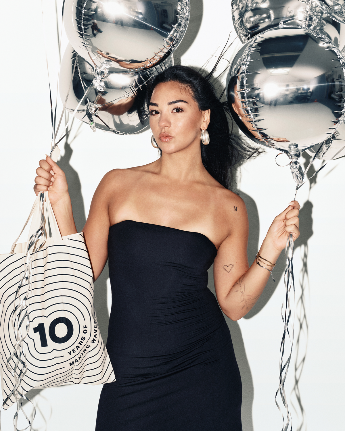 Vrouw in een strapless zwarte jurk houdt zilveren ballonnen en een gestreepte boodschappentas.
