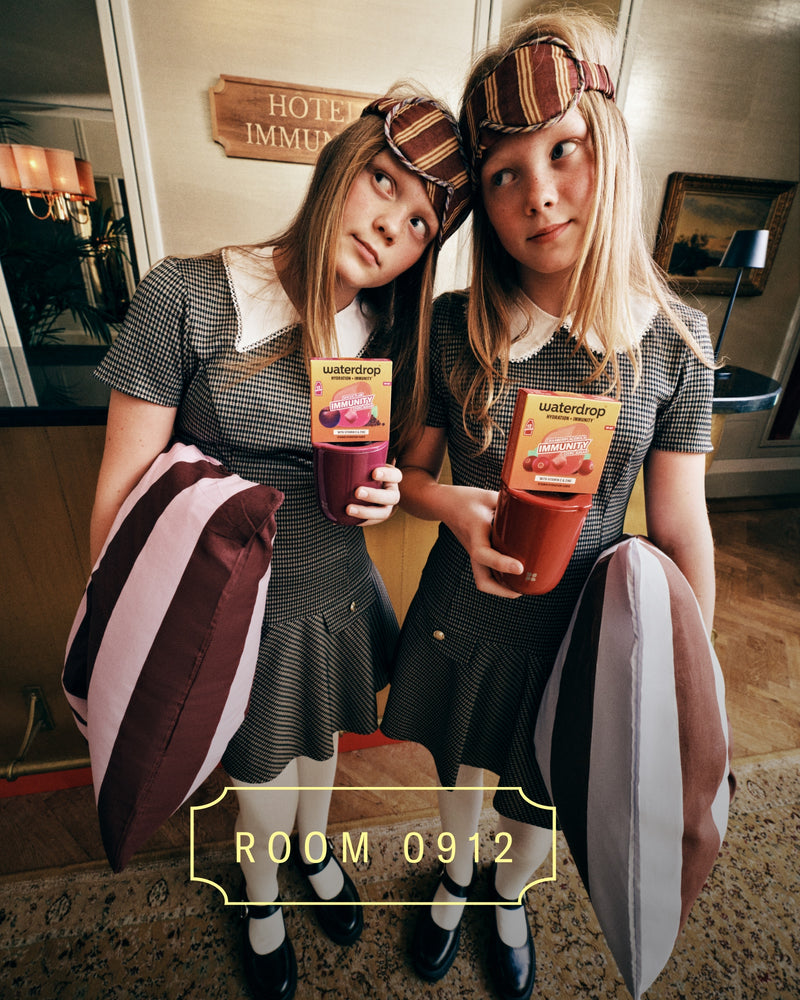 Twee meisjes in bijpassende outfits met geruite hoofdbanden houden kleurrijke drankjes in een hotelhal, staand naast elkaar met geruite tassen.