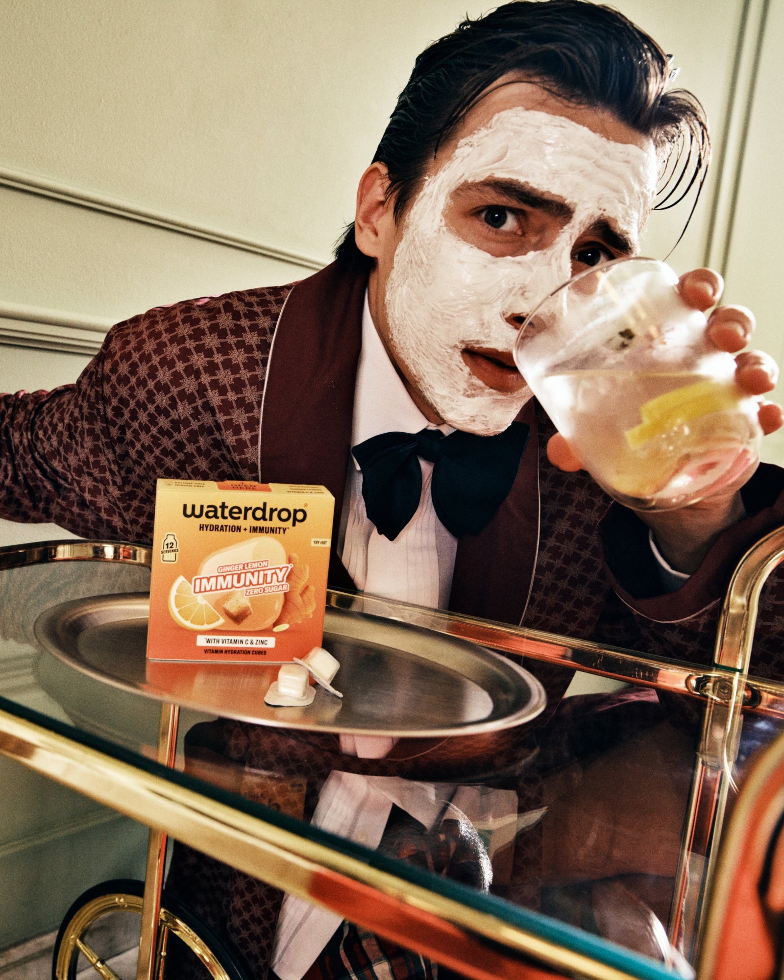 Man in een bruin pak met een gezichtsmaske drinkt uit een glas aan de bar.