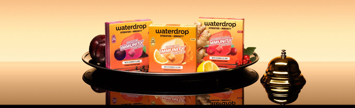 Drie waterdrop-drankkartons op een zwart dienblad met fruit op een reflecterende ondergrond tegen een warme gradientachtergrond.