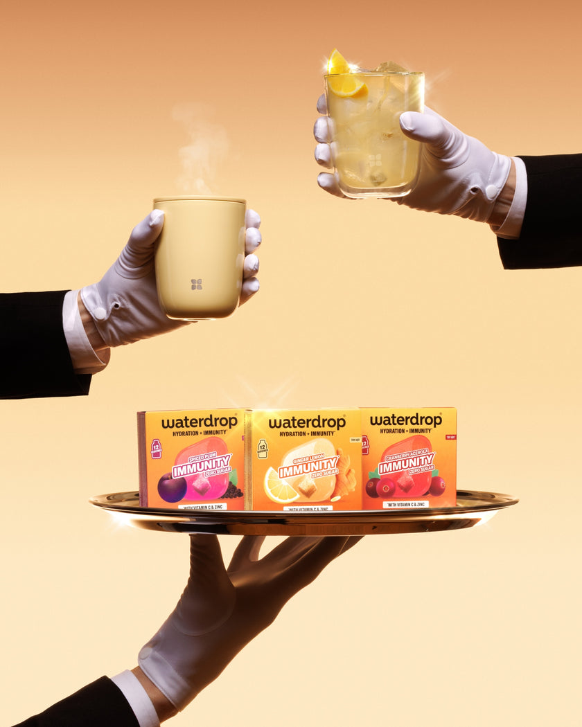 Dienblad gedragen door met handschoenen toegeruste handen met drie Waterdrop-dozen en twee kopjes, een met een schijfje citroen.