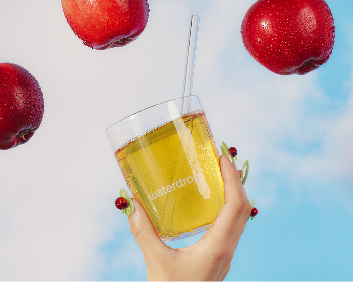 Een hand houdt een glas geel sap met een rietje vast, omringd door rode appels tegen een blauwe lucht.