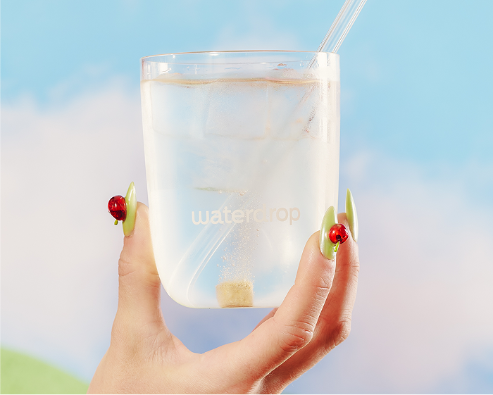 Een hand houdt een helder glas water met ijsblokjes en een rietje, tegen een blauwe lucht.