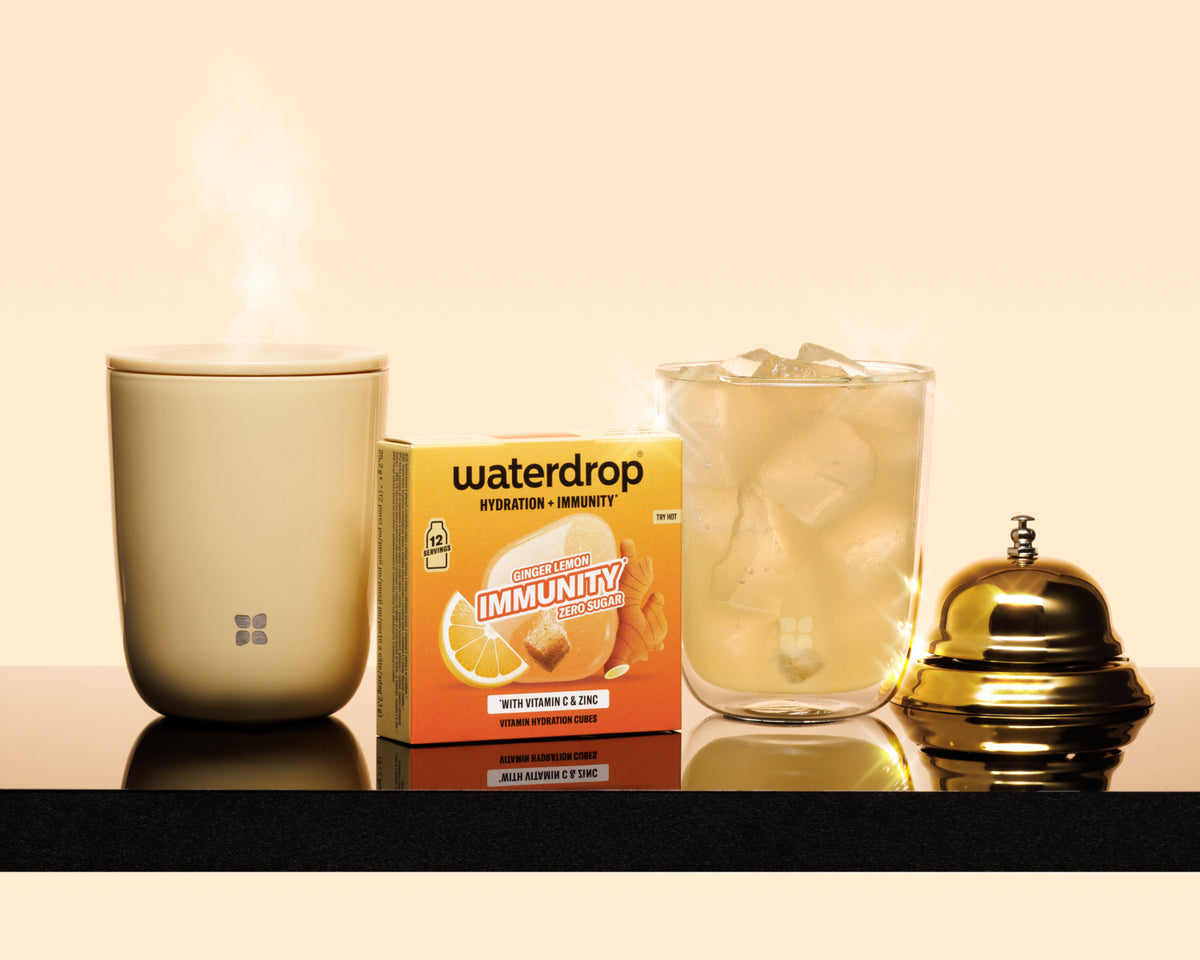 Beige dampende mok, een verpakte productdoos, een glas met ijs en een messing bel op een donker plank.