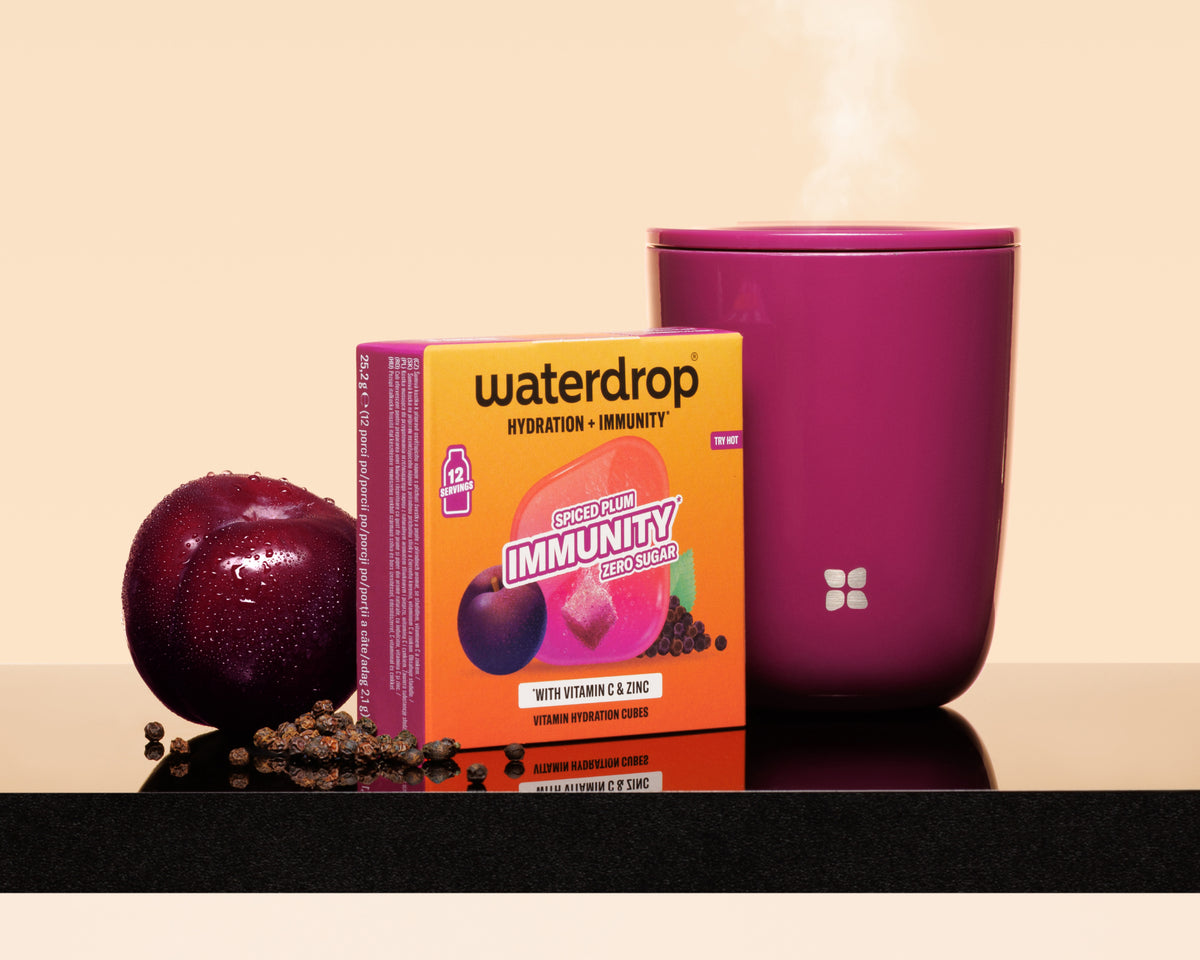 Paars kopje met stoom naast een oranje Waterdrop-doos, een pruim en verspreide zaden op een reflecterende ondergrond.