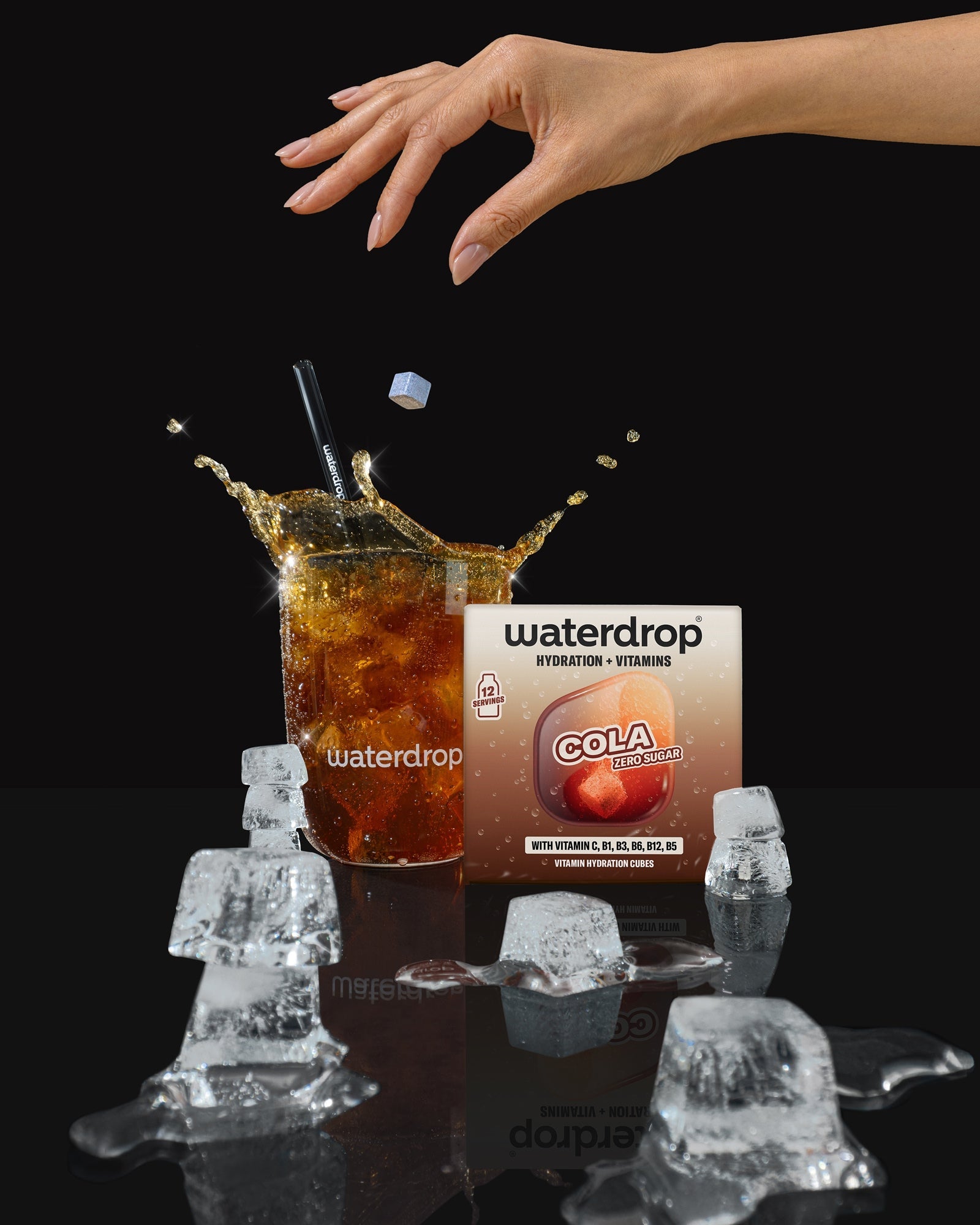 Een hand reikt over een glas cola met ijsblokjes, op de achtergrond staat een verpakkingsdoos van een drank.
