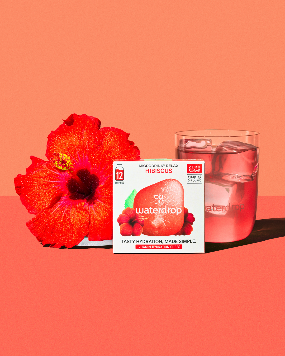 HIBISCUS Duopakket: Een doos waterdrop “Microdrink Relax Hibiscus” hydratatieblokjes met een glas roze, met ijs gekoeld water, vergezeld door een heldere rode hibiscusbloem, benadrukking op eenvoudige en smaakvolle hydratatie.