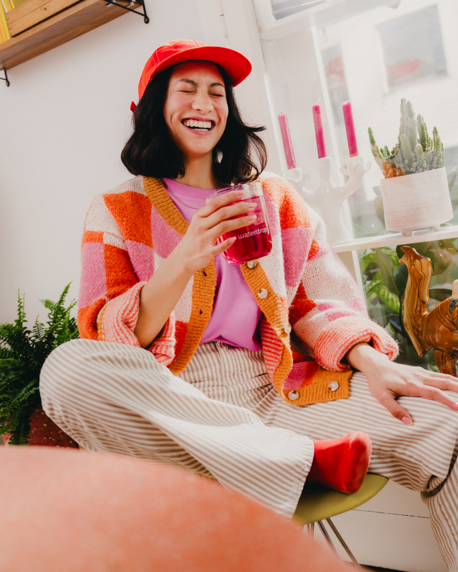 Persoon lacht terwijl ze een Microdrink Dubbelpack- korte houdbaarheid in een roze cup van waterdrop® vasthouden, zittend bij een raam met decoratieve planten en kaarsen. Geniet van deze tijdelijke aanbieding.