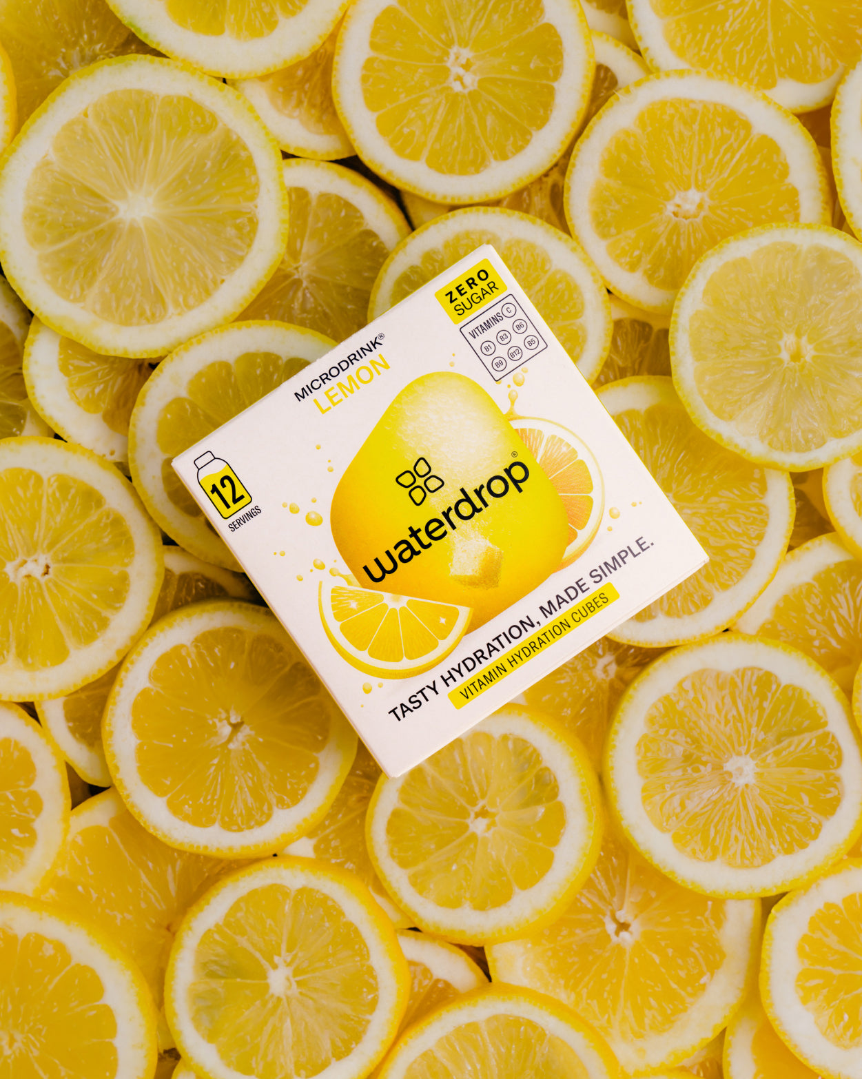 Een doos Waterdrop “Microdrink Lemon” CITROEN 3+1 vitamine hydratatieblokjes ligt op een stapel verse citroenschijfjes. De doos vermeldt “Zero Sugar” en “12 servings” voor eenvoudige, smaakvolle hydratatie.