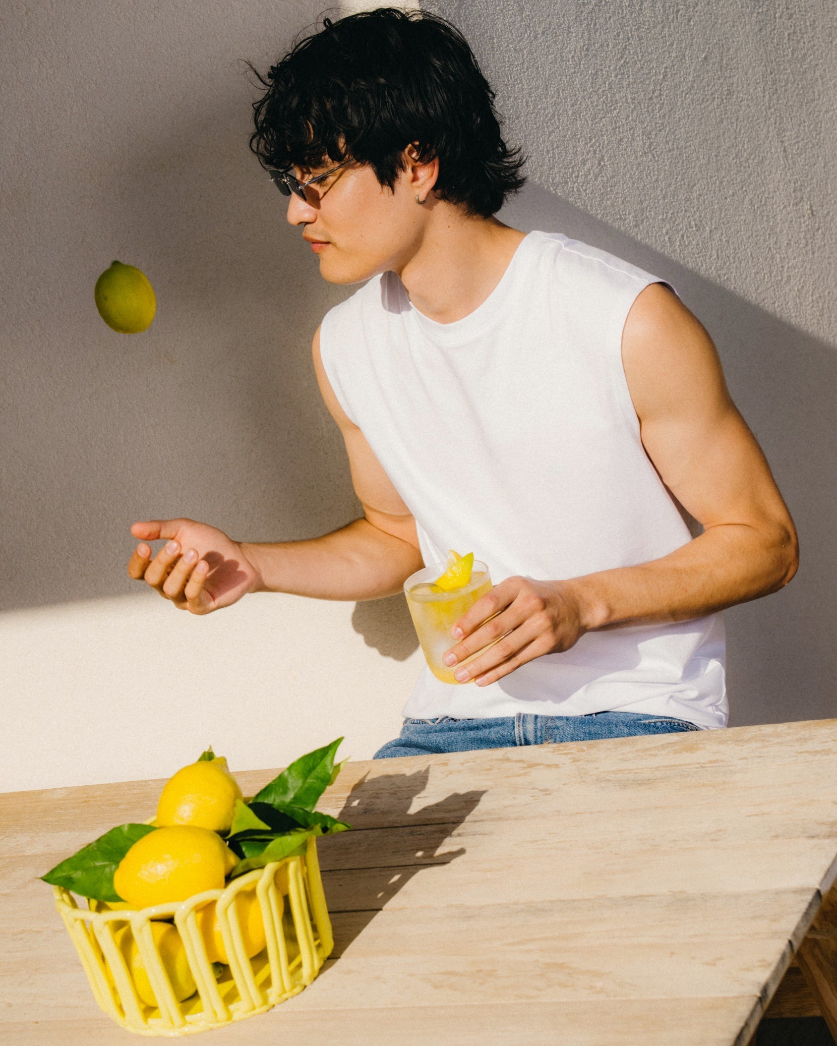 Jonge man met bril gooit citroen op, zittend aan tafel met glas citroenwater. Voor hem een mand met citroenen. Promotie voor CITROEN 3+1 van waterdrop® NL.