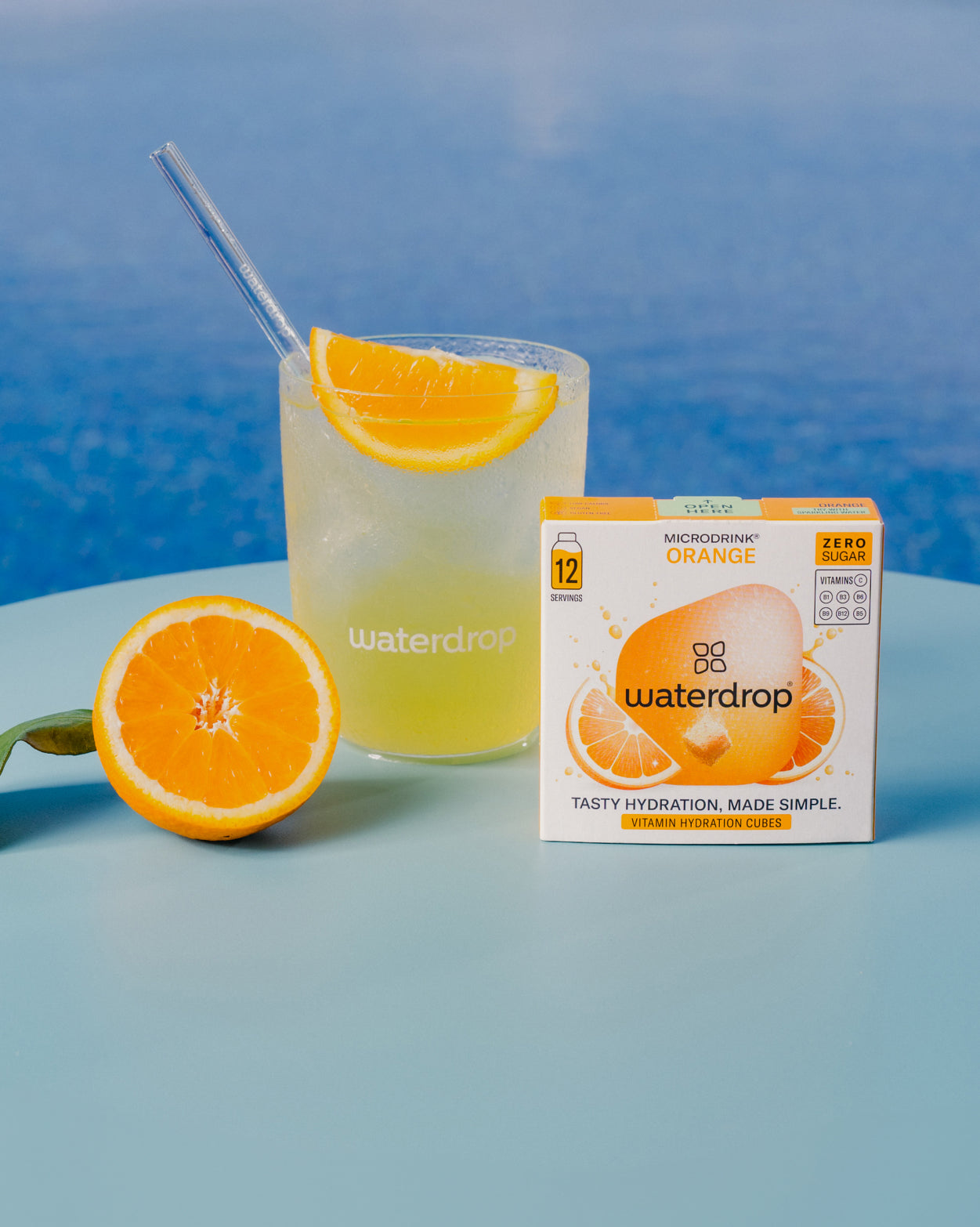 Glas met waterdrop SINAASAPPEL 2+1 SET, gevuld met verfrissend sinaasappeldrankje met ijs en herbruikbaar rietje, naast een halve sinaasappel en doos met 12 suikervrije Microdrink hydratatieblokjes.